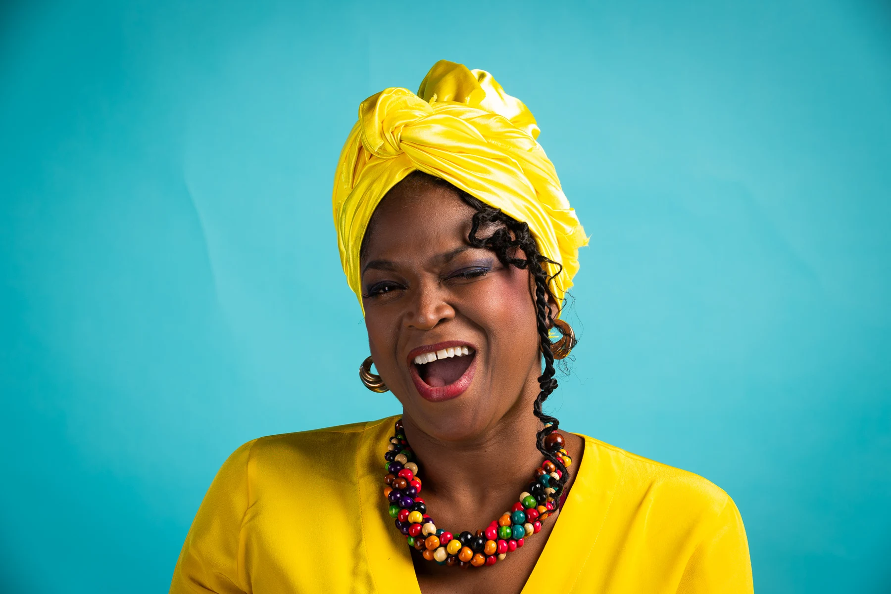 Lily sourit, en turban et chemise jaunes, sur fond turquoise